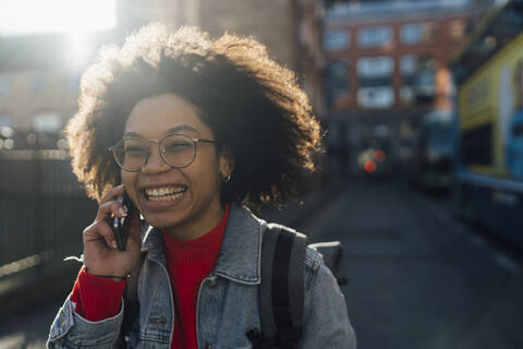 Nahaufnahme einer fröhlichen jungen Frau mit lockigem Haar, die über ein Smartphone in der Stadt spricht, lizenzfreies Stockfoto