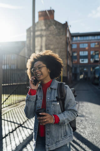 Cheerful afro woman talking over smart phone while standing with coffee cup on street stock photo