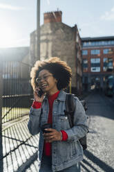 Fröhliche Afro-Frau, die über ein Smartphone spricht, während sie mit einer Kaffeetasse auf der Straße steht - BOYF01492