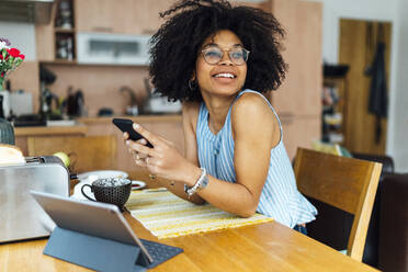 Junge Geschäftsfrau, die ein Mobiltelefon benutzt, während sie am Schreibtisch im Heimbüro sitzt - BOYF01496