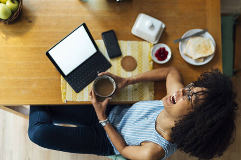 Fröhliche Geschäftsfrau mit digitalem Tablet und Frühstück auf dem Schreibtisch, zu Hause sitzend - BOYF01501