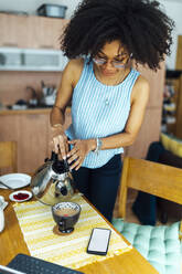 Junge Frau mit lockigem Haar gießt Wasser in Kaffeetasse auf Tisch zu Hause - BOYF01508