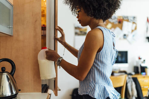 Young woman with curly hair holding milk jug while standing by refrigerator at home - BOYF01510