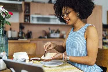 Young woman with curly hair buttering bread on table at home - BOYF01519