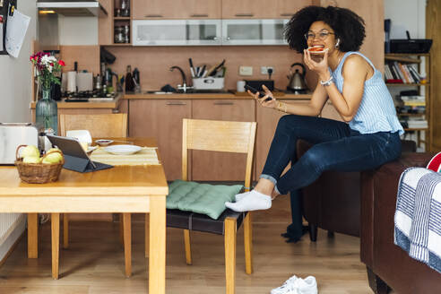 Unternehmerin mit lockigem Haar, die ihr Smartphone benutzt, während sie zu Hause Brot isst - BOYF01524