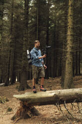 Man filming with camera and gimbal while standing on log against trees in forest - BOYF01535