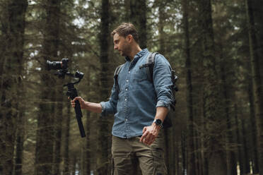 Man filming with camera and gimbal while standing against trees in forest - BOYF01536