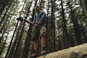 Man filming with camera and gimbal while standing on log against trees in woodland - BOYF01537