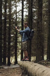 Male hiker photographing with camera while standing on log in woodland - BOYF01546