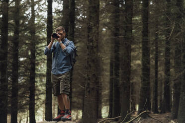 Männlicher Wanderer, der mit der Kamera fotografiert, während er an einem Baum im Wald steht - BOYF01547