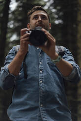 Serious male hiker holding camera looking away while standing in forest - BOYF01549