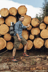 Lächelnder mittelgroßer erwachsener Mann, der auf einem Baumstamm am Holzstapel im Wald steht - BOYF01551