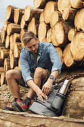 Männlicher Wanderer mit Rucksack auf einem Baumstamm sitzend gegen einen Holzstapel im Wald - BOYF01556