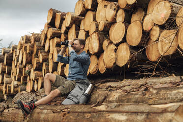 Male hiker looking through binoculars while sitting on log against woodpile in forest - BOYF01558