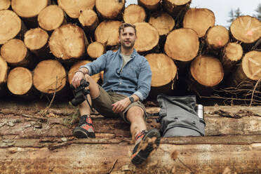 Mann mit Fernglas und Rucksack sitzt auf einem Baumstamm vor einem Holzstapel im Wald - BOYF01559