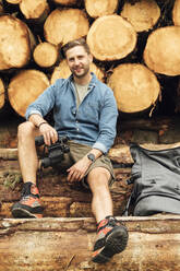 Lächelnder Mann mit Fernglas und Rucksack, der auf einem Baumstamm vor einem Holzstapel sitzt - BOYF01560