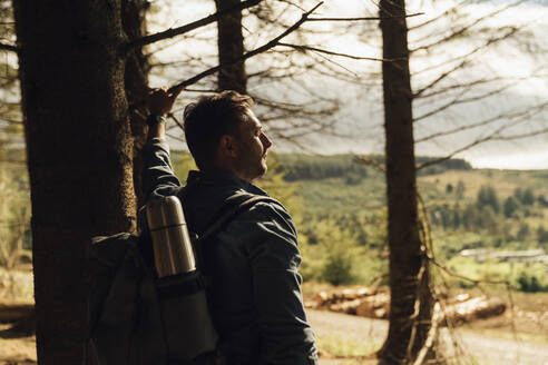 Nachdenklicher männlicher Wanderer mit Rucksack an einem Baumstamm im Wald stehend - BOYF01568