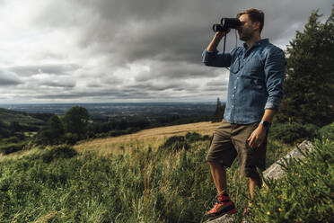 Male hiker looking through binoculars while standing on landscape against cloudy sky - BOYF01572