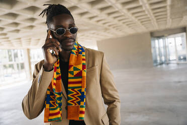 Fashionable man wearing traditional kenete talking on phone while standing in building - MPPF01071