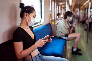 Side view of Asian female in medical mask sitting on passenger seat in train and browsing mobile phone while commuting to work during COVID 19 pandemic - ADSF14980