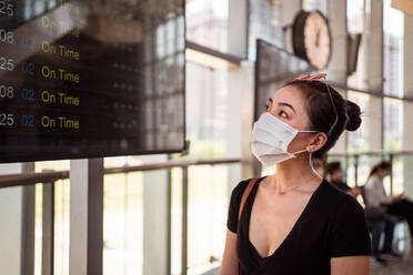 Seitenansicht einer nicht erkennbaren Frau mit medizinischer Maske, die auf einem Bahnhof steht und den Fahrplan eines Zuges während des Ausbruchs des Coronavirus betrachtet - ADSF14981