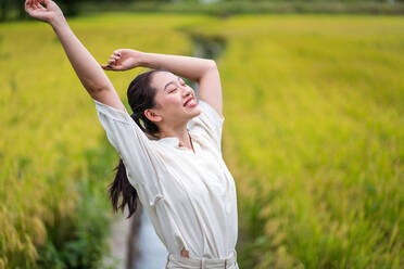 Cheerful Asian female standing in rice field with raised arms and closed eyes while enjoying freedom in summer - ADSF14985