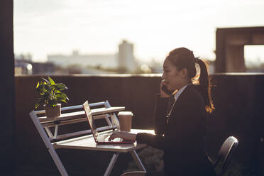 Side view of young Asian business lady in formal outfit talking on smartphone and using laptop while working remotely on rooftop terrace in city - ADSF14986