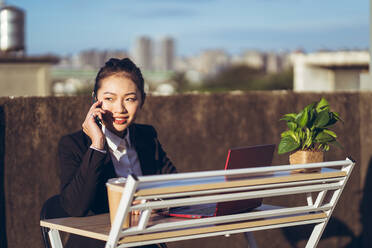 Seitenansicht einer jungen asiatischen Geschäftsfrau in formellem Outfit, die mit ihrem Smartphone spricht und einen Laptop benutzt, während sie aus der Ferne auf einer Dachterrasse in der Stadt arbeitet - ADSF14987