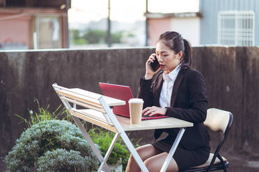 Seitenansicht einer jungen asiatischen Geschäftsfrau in formellem Outfit, die mit ihrem Smartphone spricht und einen Laptop benutzt, während sie aus der Ferne auf einer Dachterrasse in der Stadt arbeitet - ADSF14988