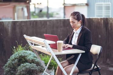 Seitenansicht einer jungen asiatischen Geschäftsfrau in formellem Outfit, die einen Laptop benutzt, während sie aus der Ferne auf einer Dachterrasse in der Stadt arbeitet - ADSF14989