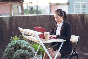 Side view of young Asian business lady in formal outfit using laptop while working remotely on rooftop terrace in city - ADSF14989