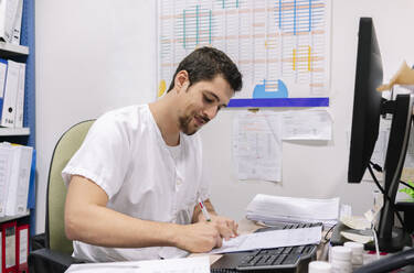 Männlicher Arzt schreibt am Schreibtisch in einer Apotheke an einem Dokument - DGOF01269