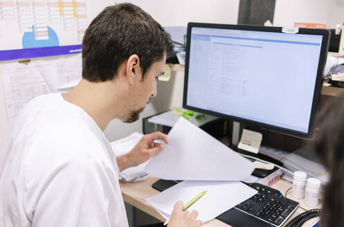 Männlicher Arzt, der am Schreibtisch sitzend in einem Krankenhaus die Krankenakte prüft - DGOF01270