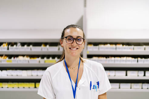 Smiling female pharmacist standing against medicine shelves in storage room - DGOF01301