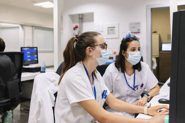 Female pharmacists wearing masks discussing over computer while sitting in hospital - DGOF01310