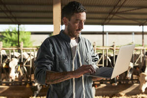 Landwirt, der einen Laptop benutzt, während er in der Nähe des Viehbestands auf dem Bauernhof steht - VEGF02873