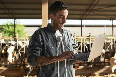 Farmer using laptop while standing near livestock at farm - VEGF02873