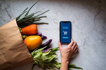 Anonyme Frau an einem Tisch mit verschiedenen Obst- und Gemüsesorten, die ein Smartphone benutzt und Essen über einen Online-Lieferdienst bestellt, aus einem hohen Winkel - ADSF15004