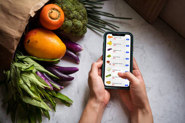 Draufsicht auf eine gesichtslose Frau am Tisch mit Papiertüte mit frischen Lebensmitteln, die Lebensmittel auswählt und über eine App auf dem Smartphone online einkauft - ADSF15006