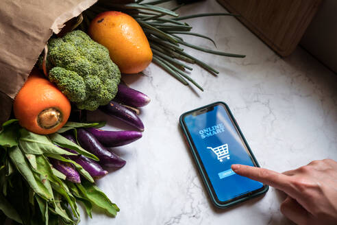 High angle of anonymous female at table with various fruits and vegetables using smartphone and ordering food via online delivery service - ADSF15008