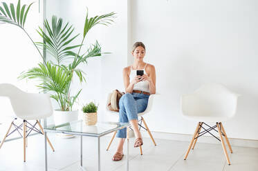 Full body female client sitting on chair and browsing on smartphone while waiting for appointment in lobby of modern clinic - ADSF15036