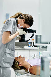 Seitenansicht einer Zahnärztin mit Handschuhen und Maske, die die Zähne einer Frau während der Arbeit in einer modernen Klinik untersucht - ADSF15039