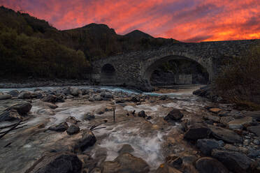 Fluss und steiniges Ufer, das durch Felsen und eine alte Steinbrücke bei Sonnenuntergang fließt - ADSF15054