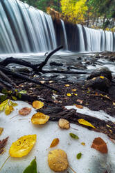 Malerische Szenerie eines reißenden Flusses mit breitem Wasserfall und felsigem Grund, der durch einen Herbstwald mit bunten Bäumen an einem bewölkten Tag fließt - ADSF15056