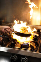 Abgeschnittener, nicht erkennbarer Koch beim Braten von Garnelen in der Flamme über dem Gasherd mit brennender Flamme beim Kochen im Restaurant - ADSF15101
