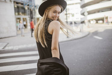 Smiling woman looking over shoulder while standing on street - KMKF01472