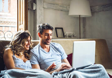 Glückliches junges Paar sitzt zu Hause auf der Couch und schaut auf den Laptop - EHF00920