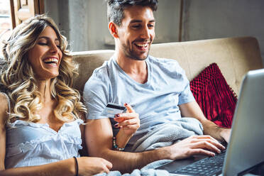 Happy young couple sitting on couch at home with credit card and laptop - EHF00923