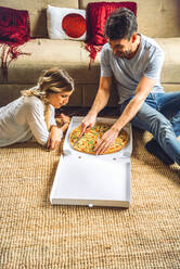 Junges Paar sitzt auf dem Boden des Wohnzimmers und schneidet eine Pizza im Karton - EHF00946