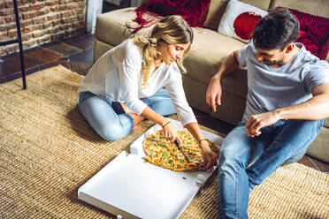 Junges Paar sitzt auf dem Boden des Wohnzimmers und schneidet eine Pizza im Karton - EHF00947